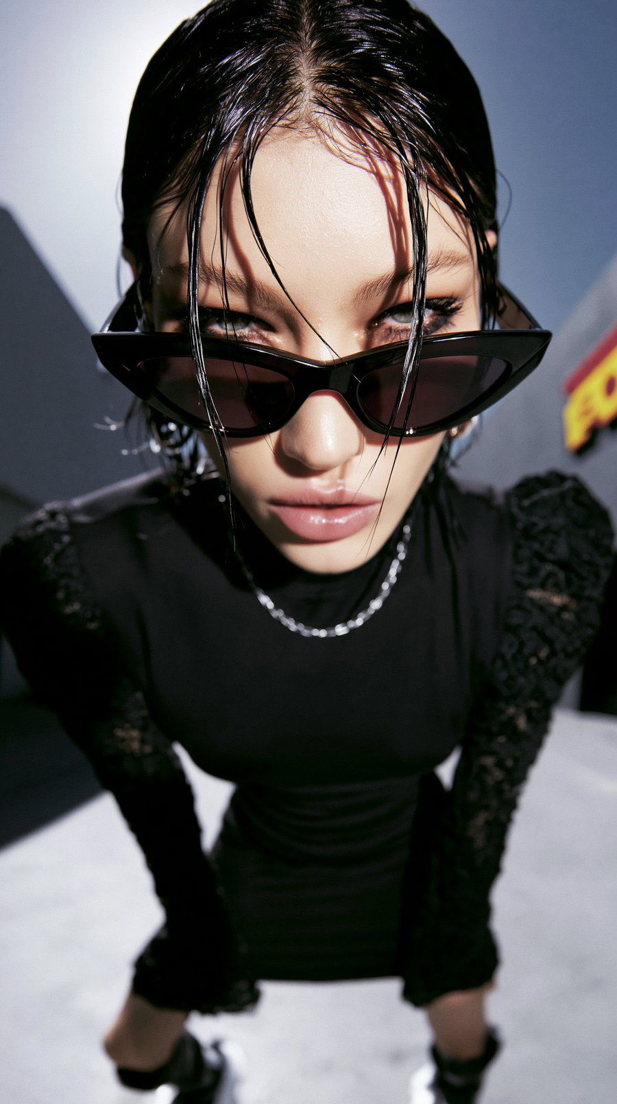 Close-up image of a woman with wet hair wearing black cat-eye sunglasses, a black dress with lace sleeves, and a silver chain necklace, looking directly at the camera