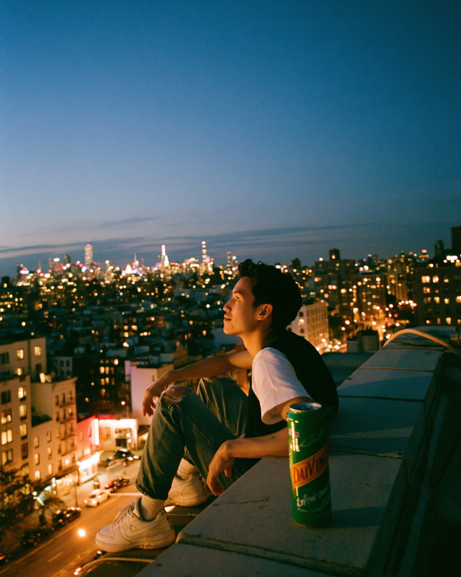 A young man in casual clothes sitting on a rooftop ledge with city lights and buildings in the background during dusk. A can of DaVinci soft drink is placed on the ledge nearby.