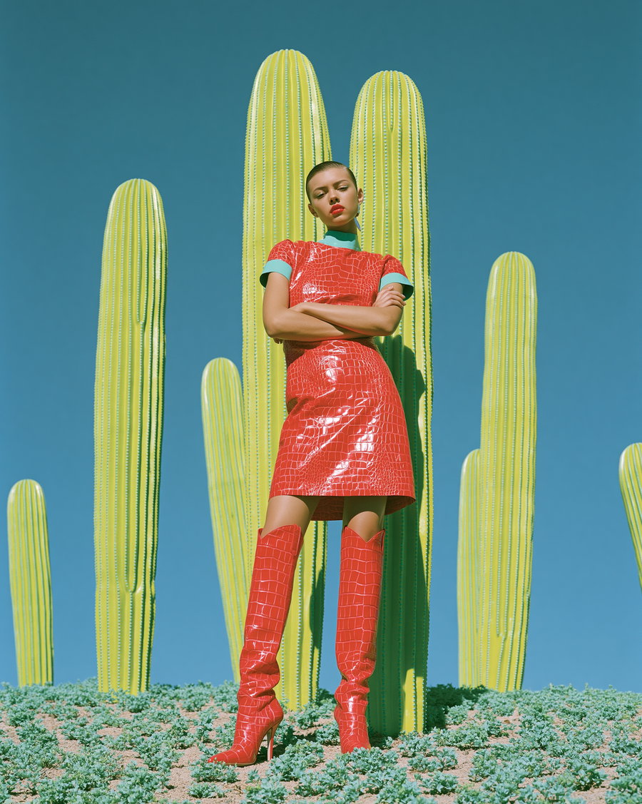 A woman in a shiny red crocodile-pattern short dress and matching knee-high boots stands confidently with arms crossed in front of tall, yellow cacti under a clear blue sky