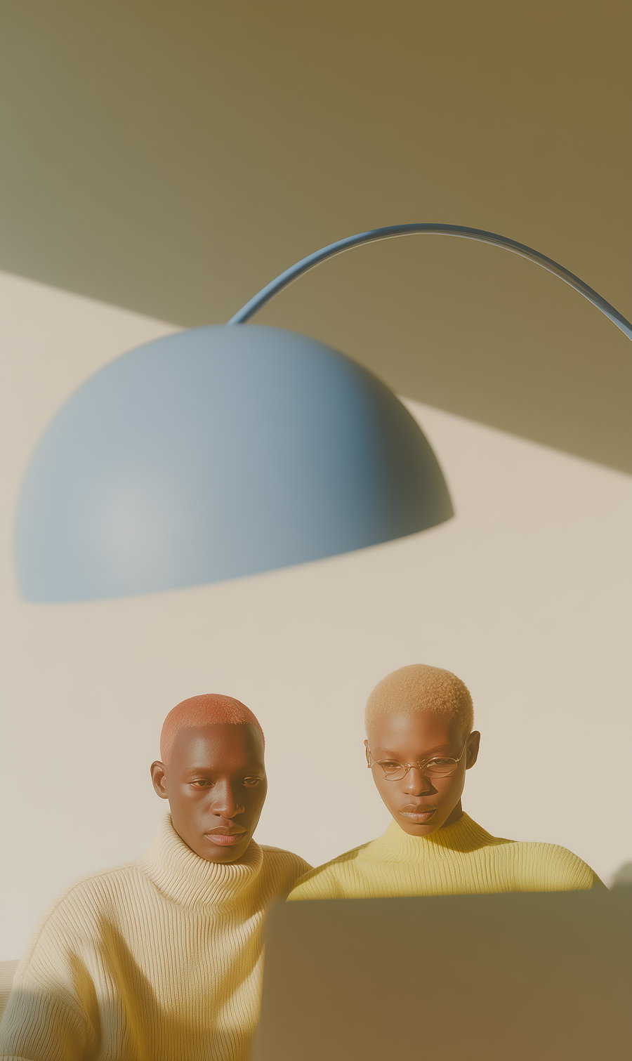 Two individuals with short colored hair and glasses wearing cozy sweaters, sitting together and focused on something in front of them, illuminated by soft natural light and a large modern light fixture above.