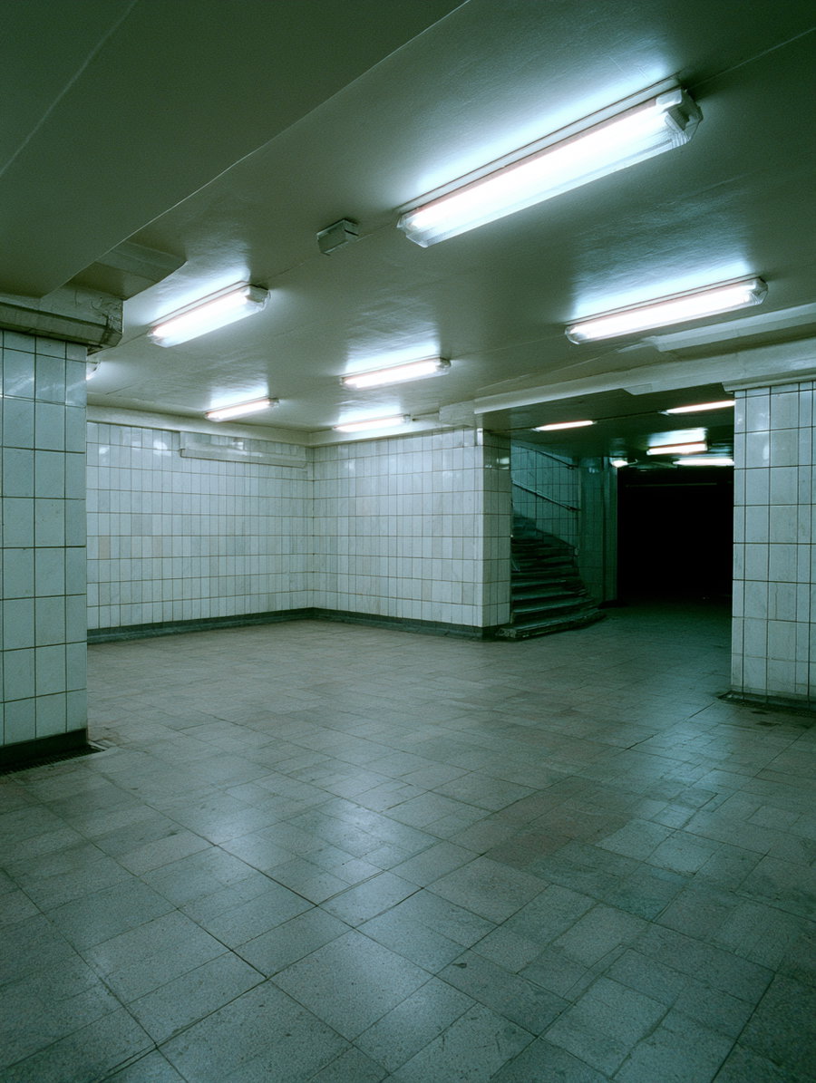 Empty underground corridor with white tiled walls, fluorescent ceiling lights, and a stairway leading upwards in the background