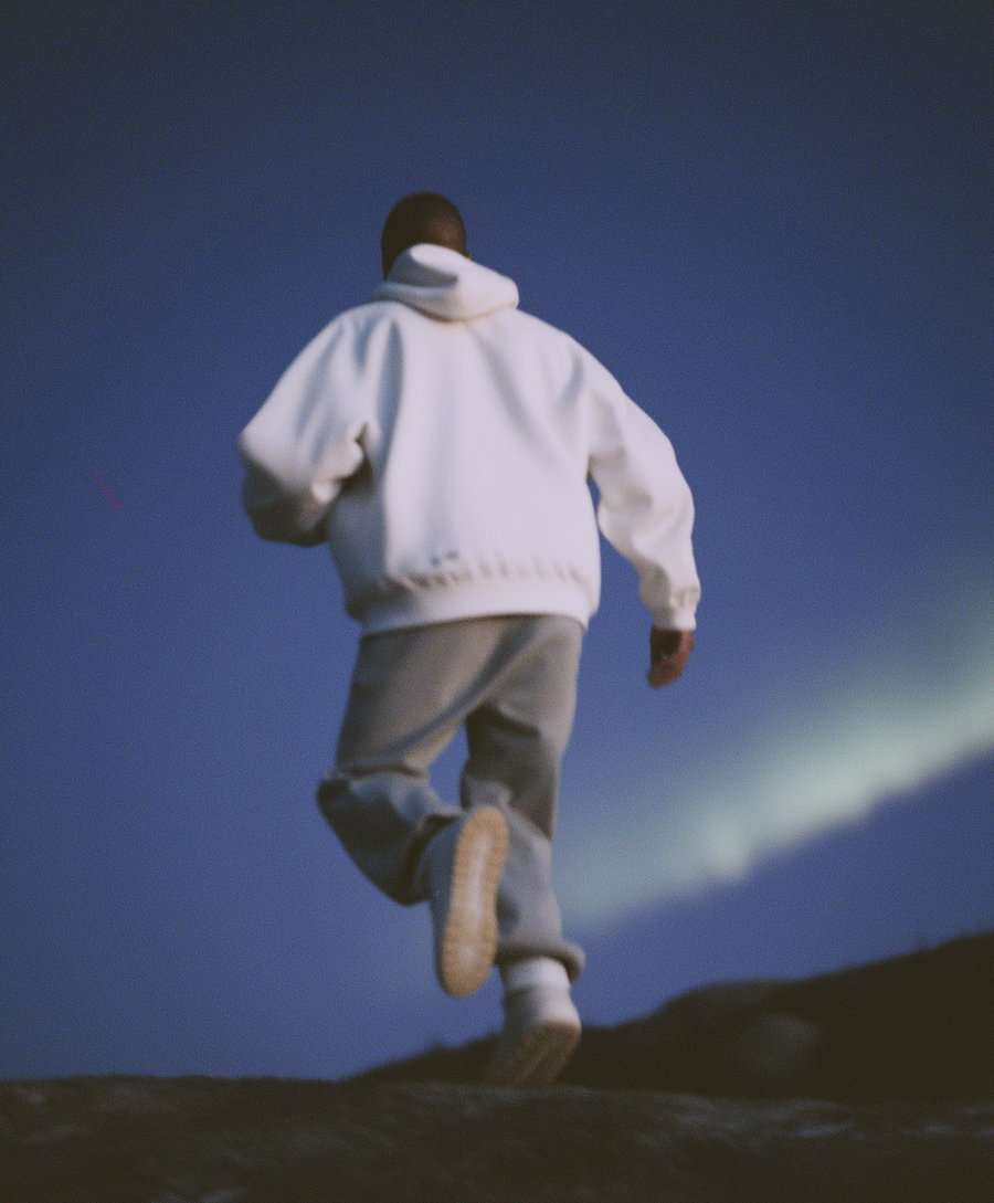 A person wearing a white hoodie and gray pants walking on rocky terrain under a dark blue sky with a faint light in the background.