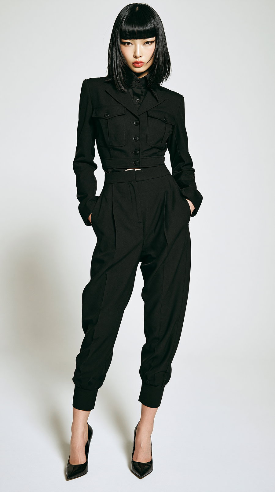 A woman wearing a black structured jacket, matching high-waisted pants, and black high heels standing against a plain light backdrop.