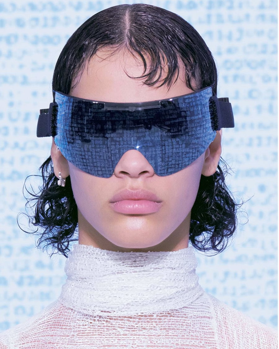 Close-up portrait of a person with wet-look dark hair wearing large futuristic blue visor-style eyewear and a high-neck white textured top against a blue patterned background