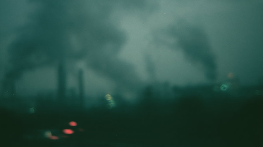 Blurry and dark photo of industrial smokestacks emitting dense smoke into a moody, foggy sky