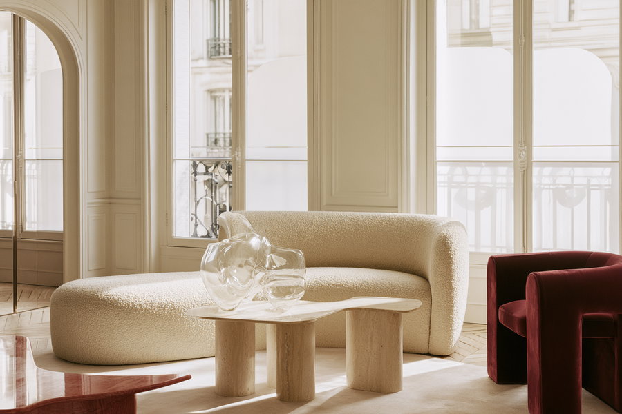 A stylish living room featuring a curved light-colored sofa, two red armchairs, and a minimalistic table beside a glass sculpture, illuminated by natural light through large windows.