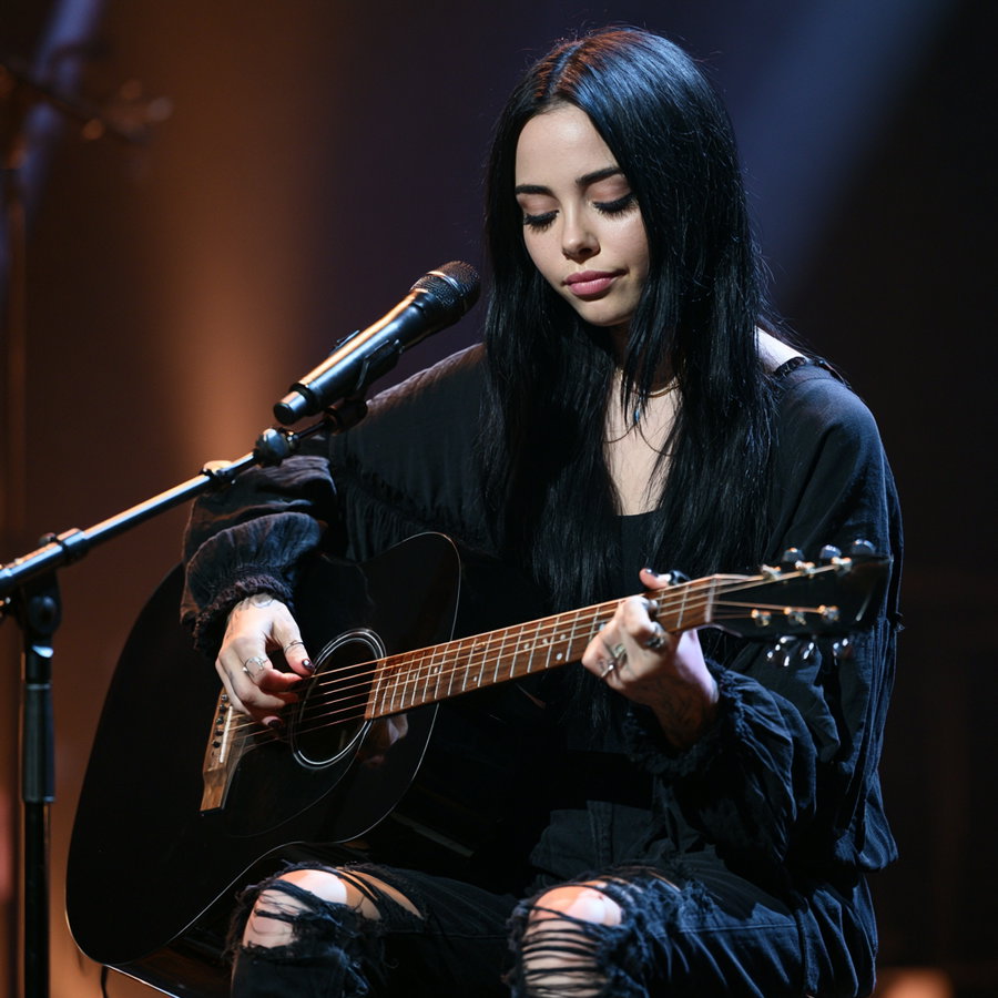 A young woman with long black hair wearing a black outfit plays a black acoustic guitar while sitting and singing into a microphone under stage lighting