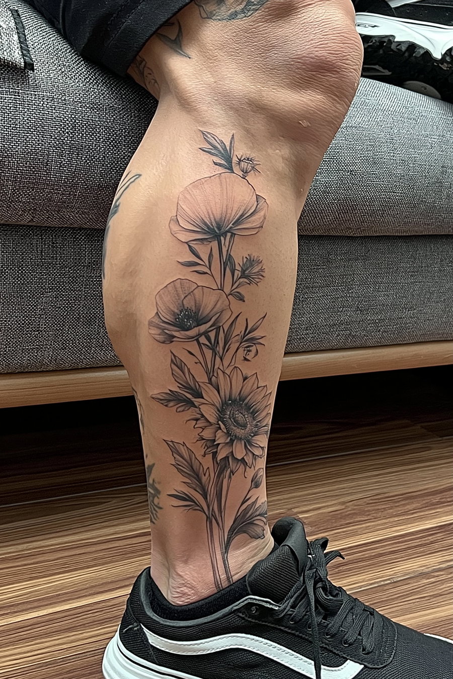 close-up of a lower leg with a black and white tattoo featuring detailed flowers and leaves, person wearing black and white sneakers, wooden floor and grey couch in background