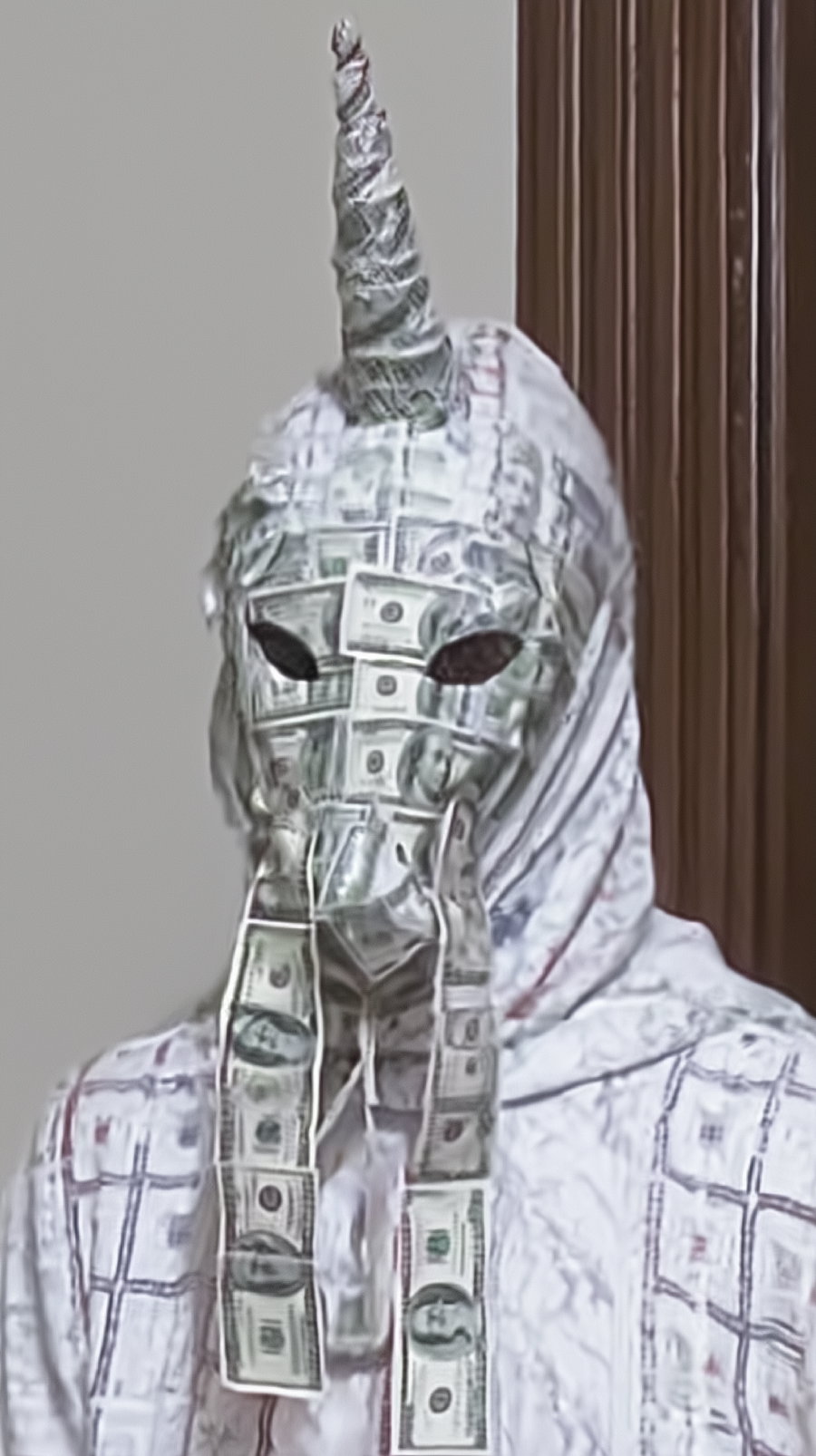 Person wearing a detailed mask covered in dollar bill patterns with a twisted unicorn horn on top, dressed in a white hooded garment with faint patterns