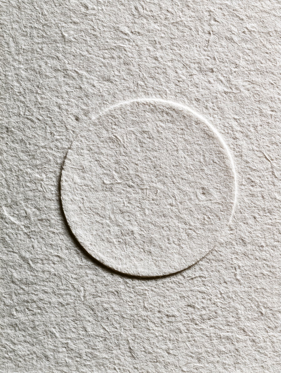 A close-up of textured white paper featuring a subtle circular embossed pattern in the center.