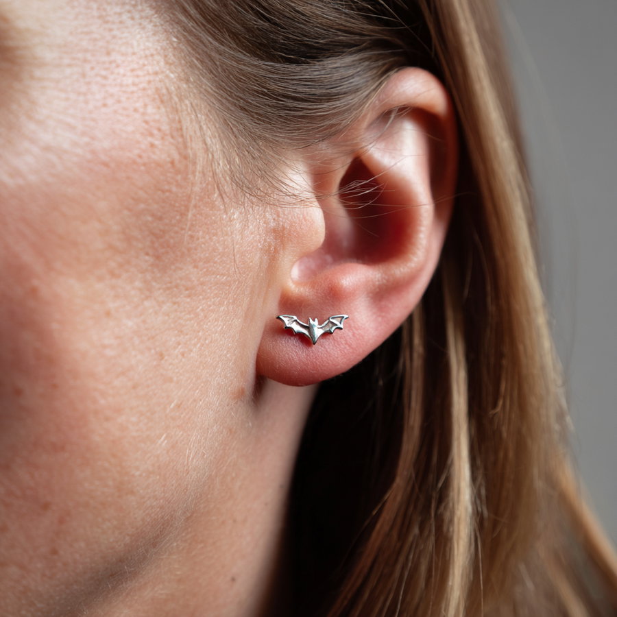 A close-up image showing a silver stud earring shaped like a bat on a person's ear