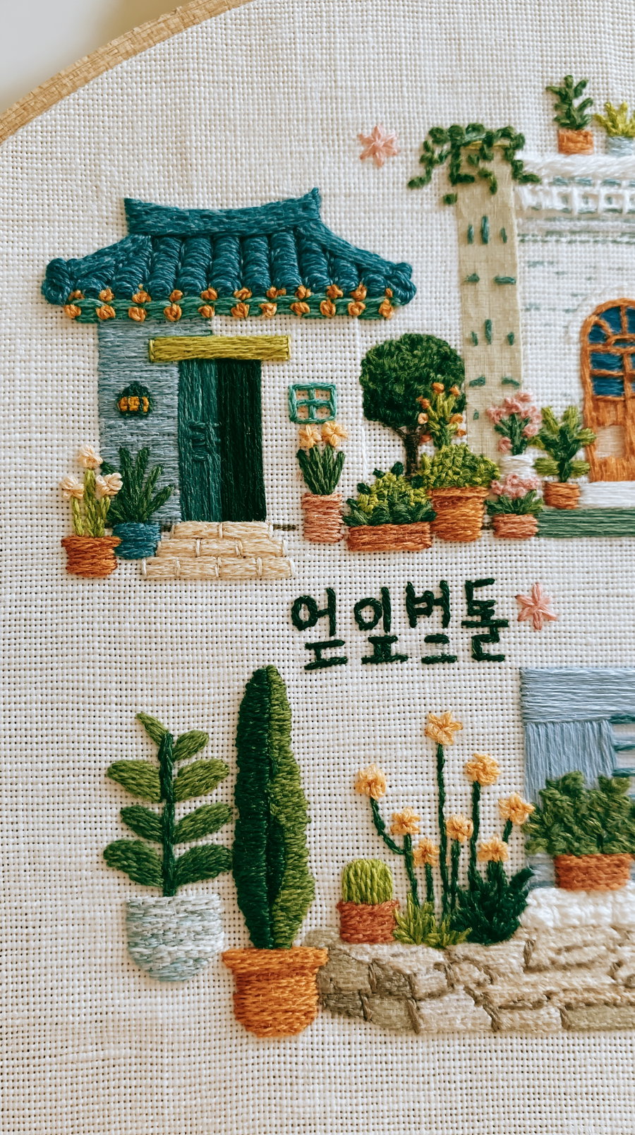Close-up embroidery showing various potted plants, a green door with a blue roof house, and Korean text on a white woven fabric