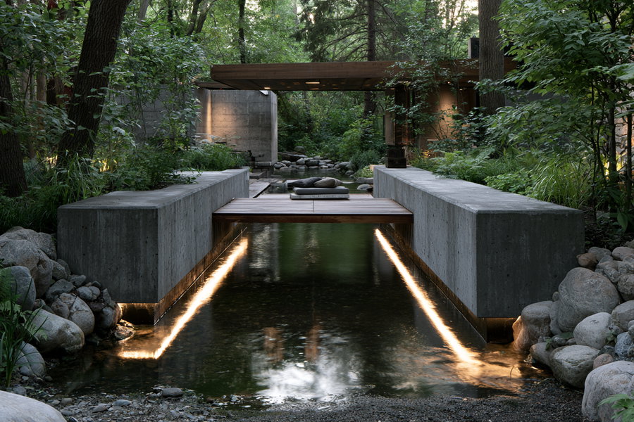 Modern outdoor seating area with concrete benches on either side of a small water stream with soft lighting beneath the benches, surrounded by dense trees and rocks