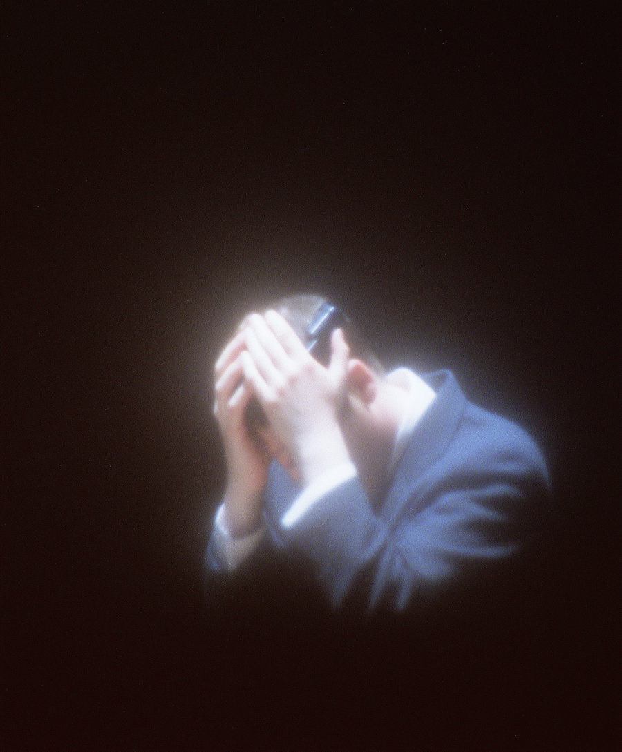 Blurred image of a person in a suit holding their head with both hands against a dark background