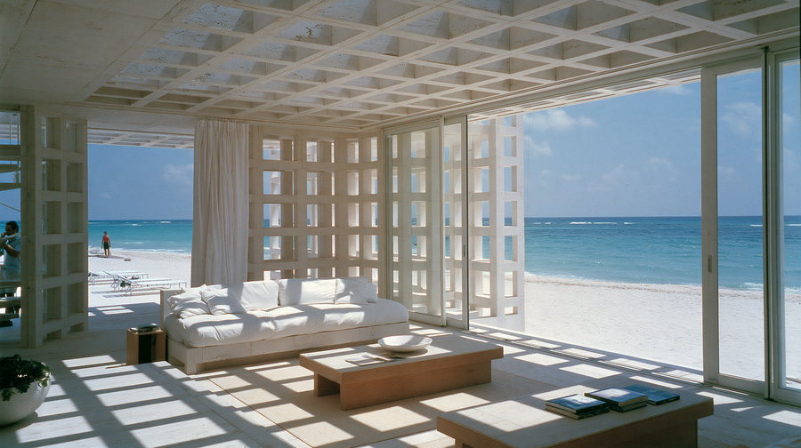 Bright living room with white sofa, wooden coffee table, lattice white walls, and large glass sliding doors overlooking sandy beach and blue ocean