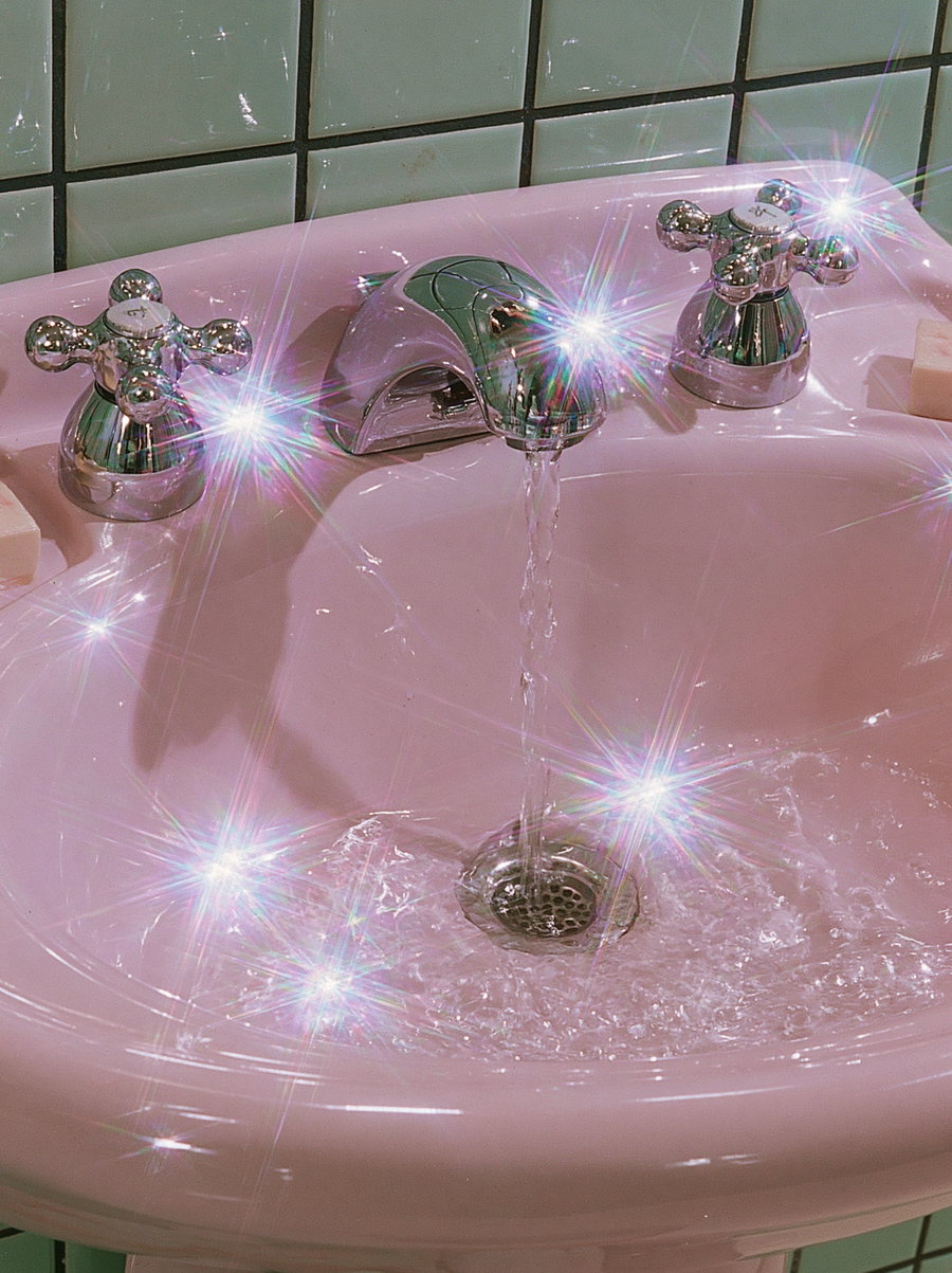 Close-up of a pink ceramic bathroom sink with chrome faucets running water and sparkles throughout the image
