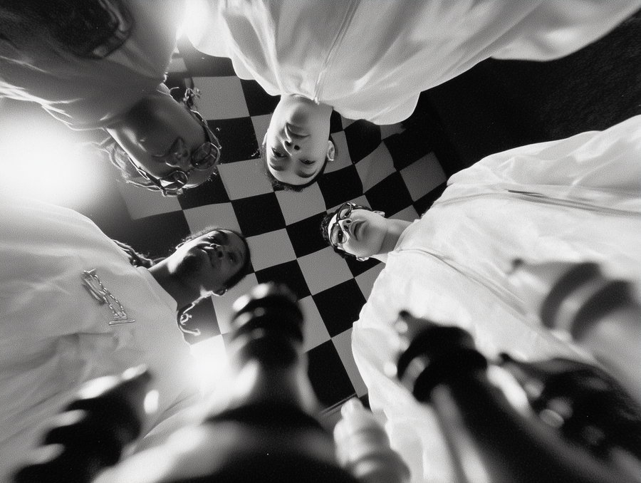 Four individuals in white coats are looking down at the camera with a blurred chessboard in the foreground, set against a checkered black and white wall background