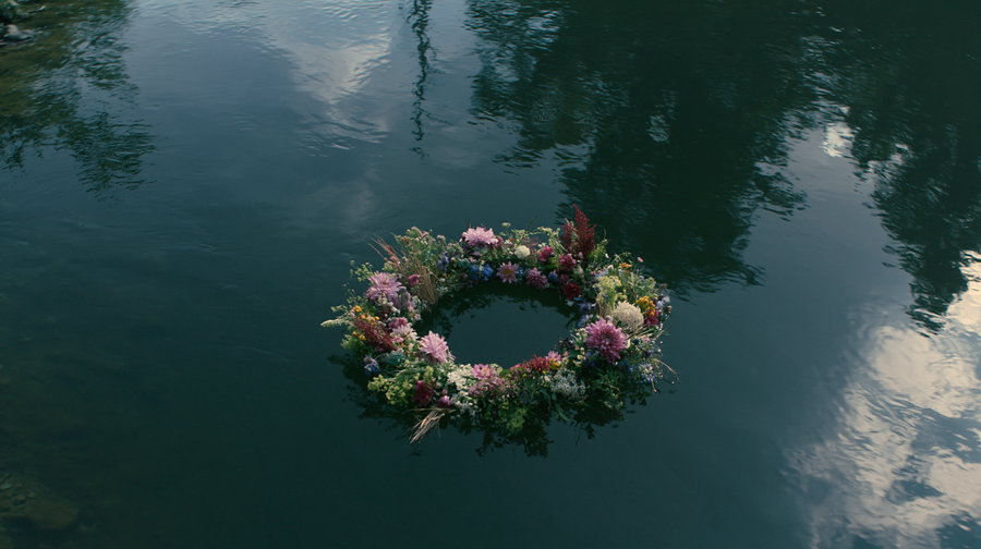 Floral wreath made of pink, purple, white, and green flowers and plants floating on calm dark water reflecting sky and trees