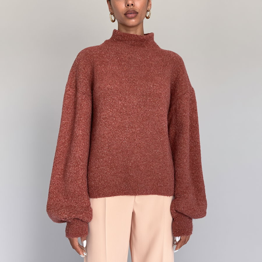 person wearing a rust-colored knitted sweater with balloon sleeves and light beige pants against a plain background