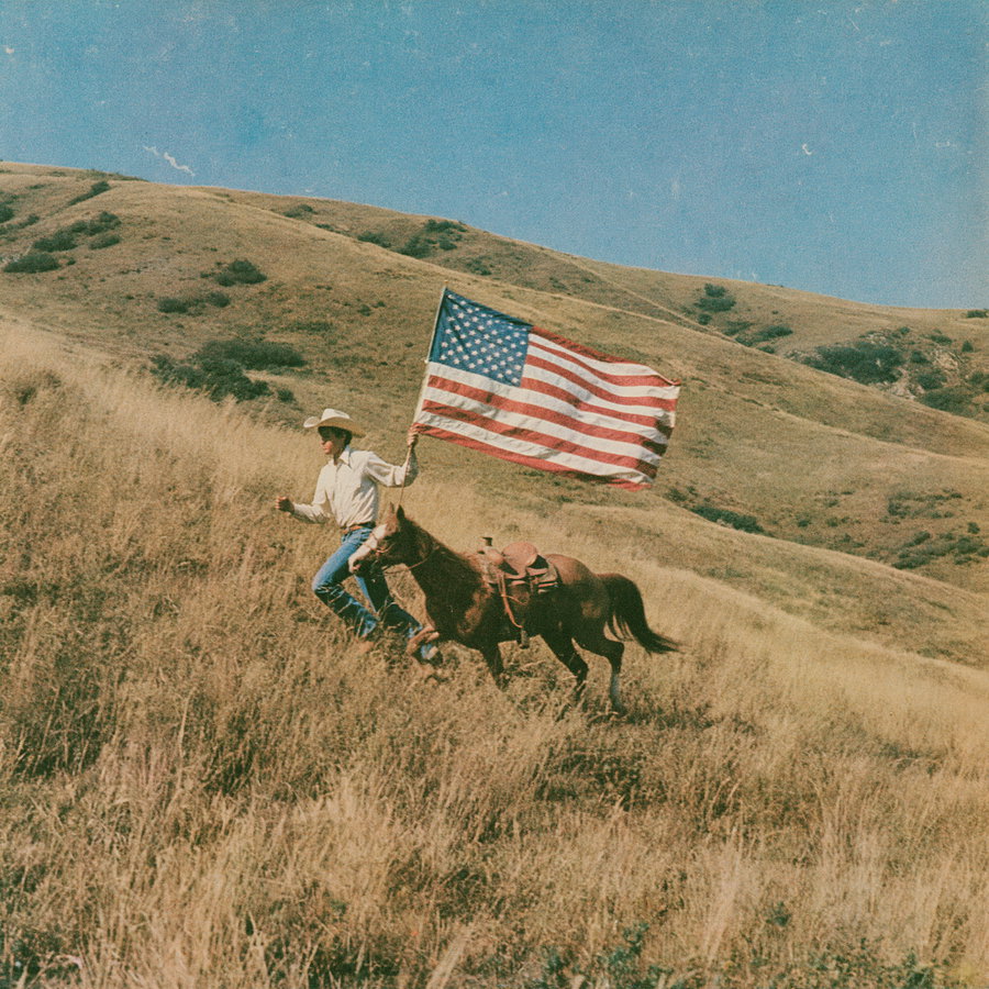 A person wearing a cowboy hat and white shirt running up a dry grassy hill while holding an American flag, with a horse beside them under a blue sky