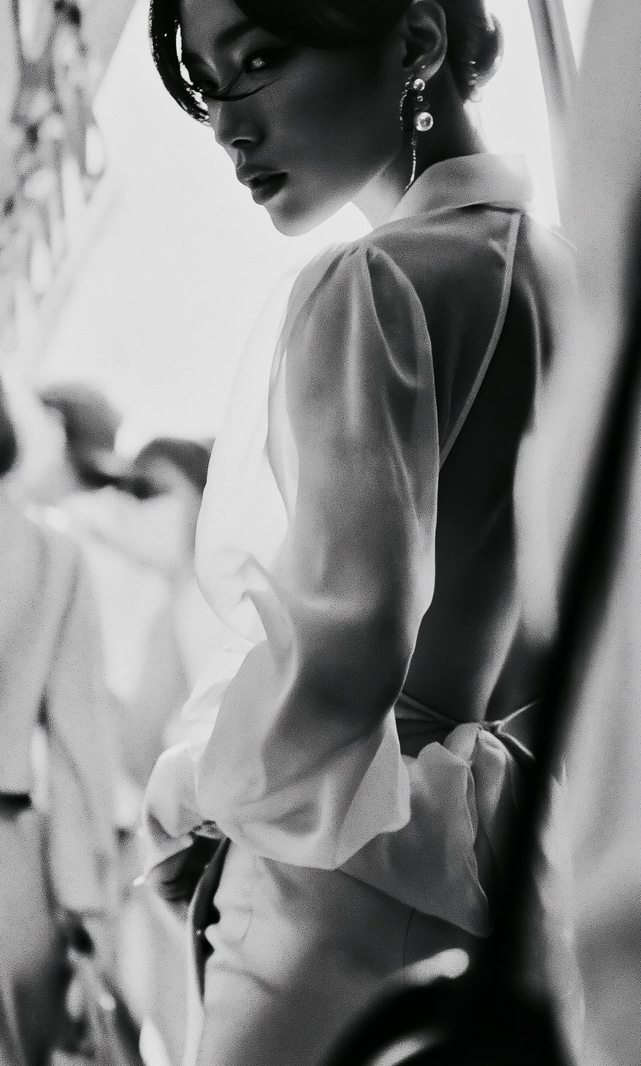 Black and white photo of a woman in a sheer, long-sleeved dress with puffed sleeves, wearing dangly earrings and looking over her shoulder.