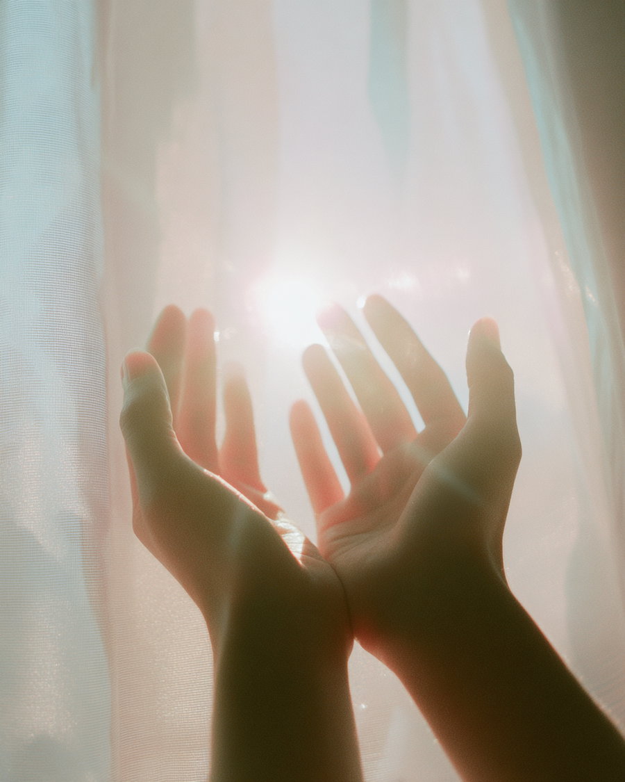 Two hands held up towards soft glowing sunlight filtered by white sheer curtains
