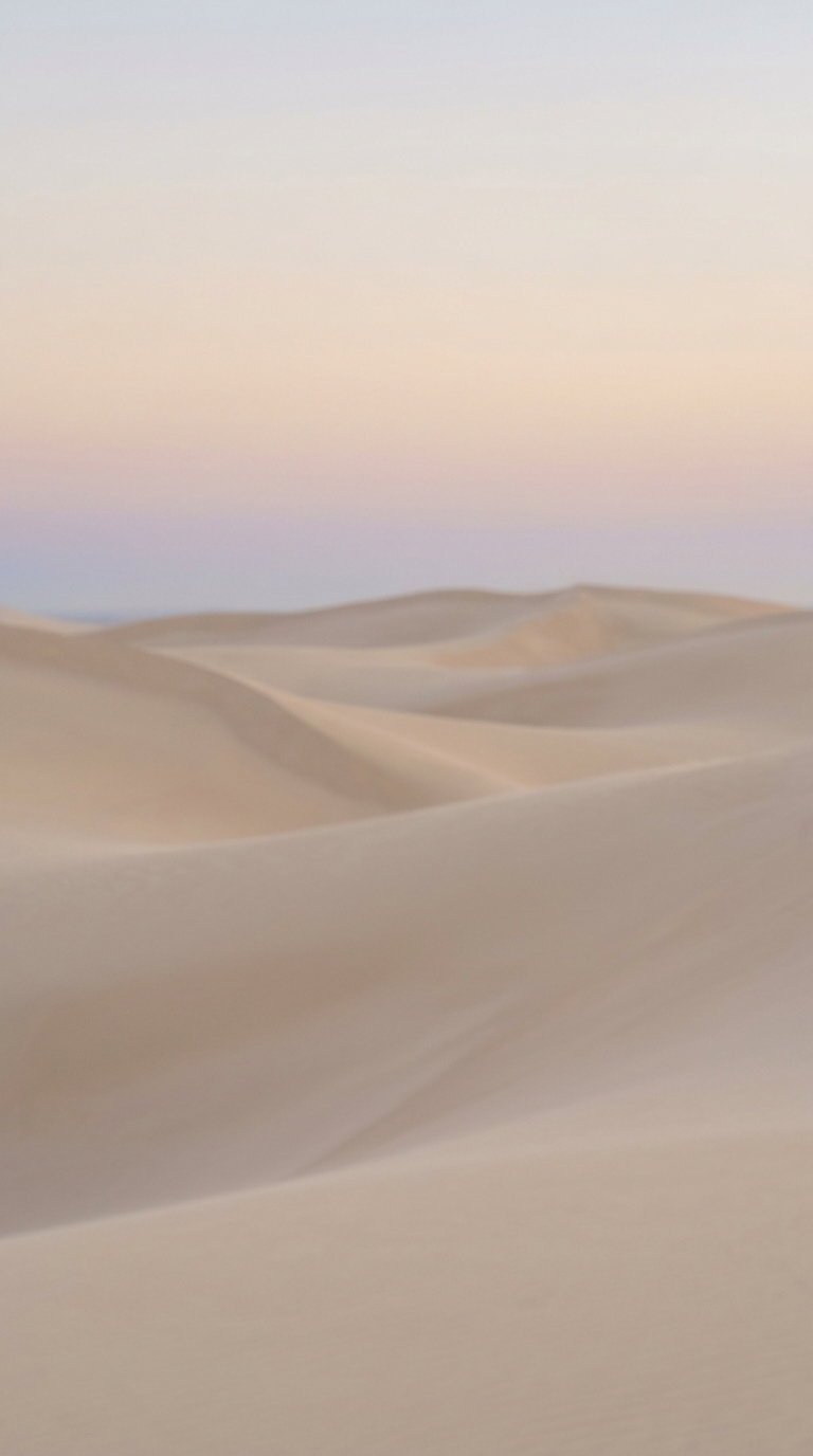 View of smooth sand dunes under a gradient pastel sky with soft hues of pink, purple and beige