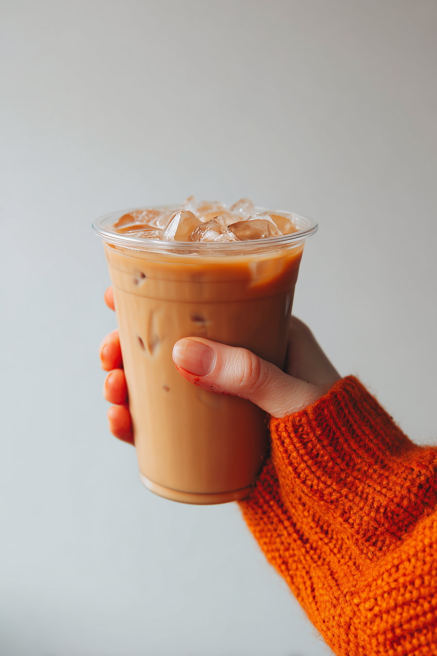 A hand wearing an orange knitted sweater holding a clear plastic cup filled with iced coffee and ice cubes against a plain neutral background