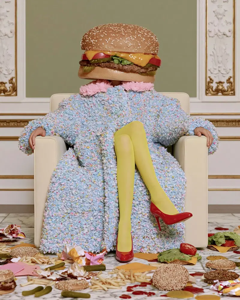 Surreal photo of person with hamburger head sitting among fast food items
