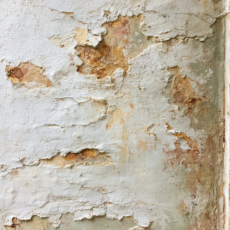 A wall surface with cracked and peeling paint in shades of white, beige, and brown revealing layers underneath