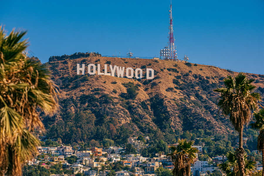 View of the Hollywood sign on a hillside with dry vegetation, communication antennas, trees, residential houses, and palm trees framing the scene