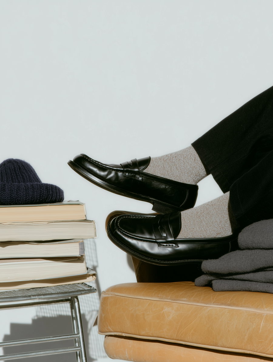 Person wearing black leather loafers with beige socks sitting on a light brown leather seat, next to a metal stand with a stack of books and navy knit hat