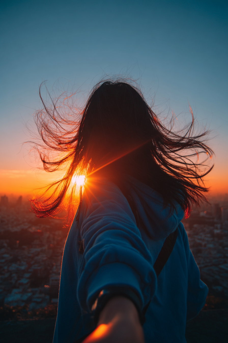 Silhouette of a person with long hair blowing in the wind, reaching out hand, with a cityscape and vibrant orange and blue sunset in the background