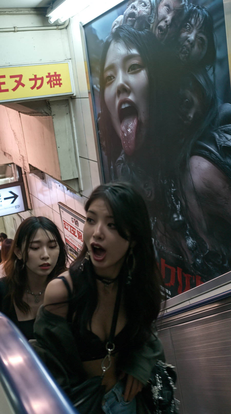 Photo of two women on an escalator in a subway with a large, dark modern poster of a woman with multiple faces and tongue out on the wall