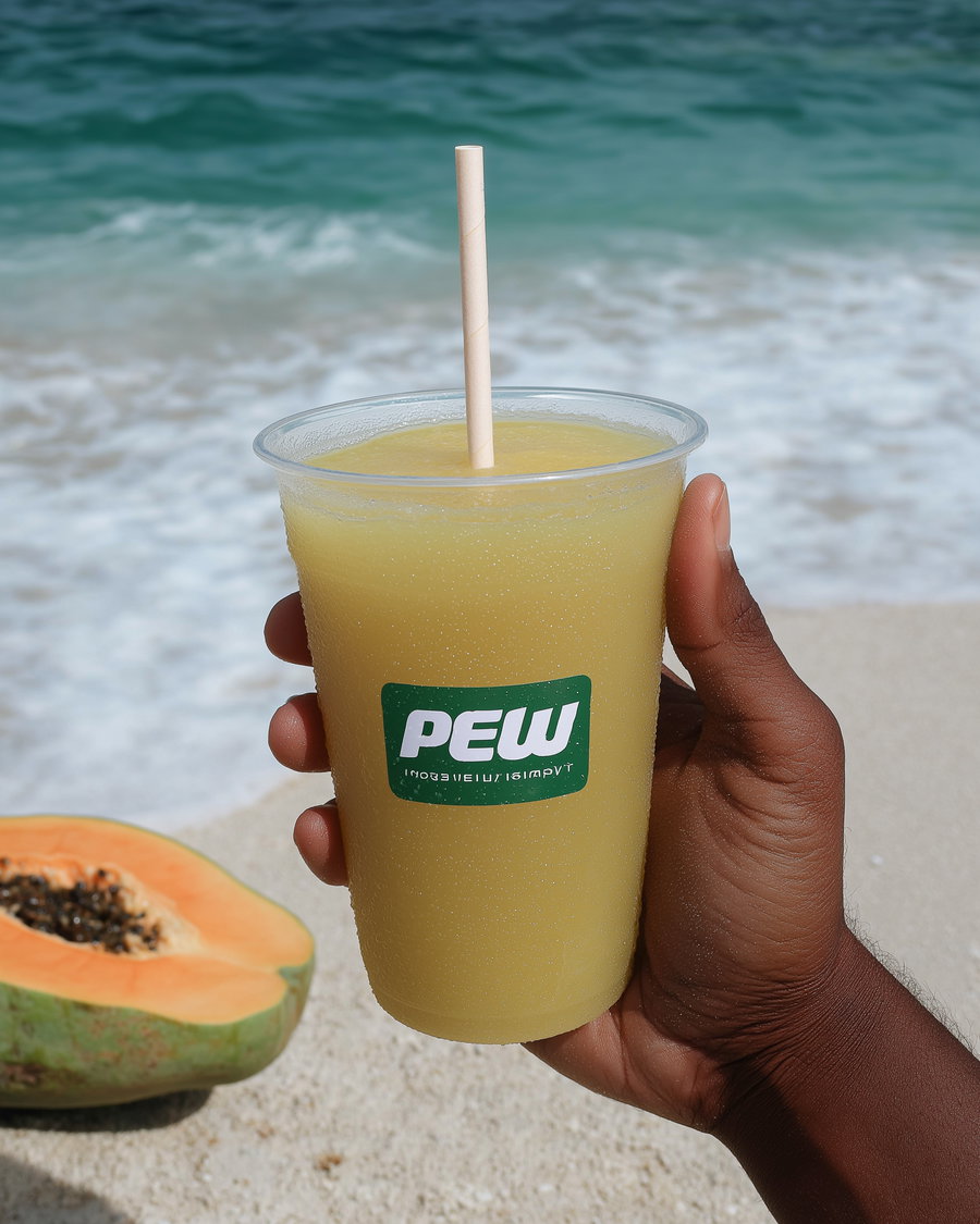 Hand holding a plastic cup filled with a yellow slushy drink with a paper straw on a beach with waves and a papaya cut in half nearby