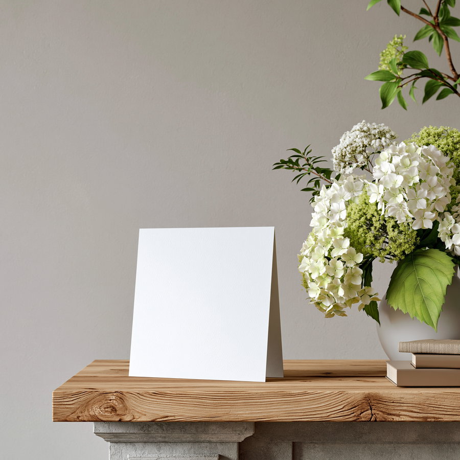 A blank greeting card standing upright on a wooden mantelpiece with a bouquet of white and green flowers in a vase nearby.