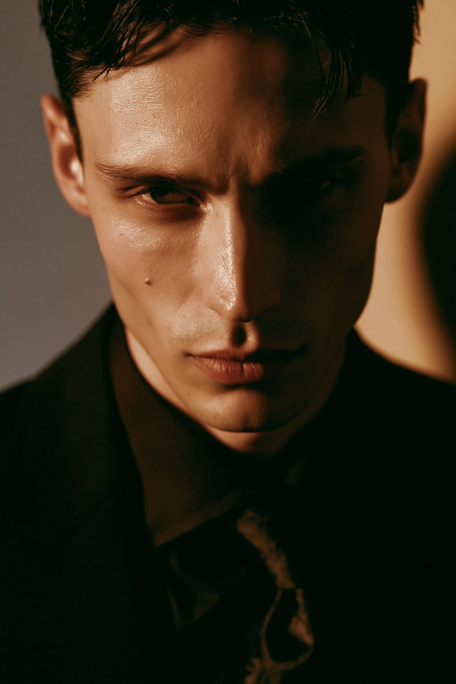 A cinematic close-up fashion portrait of a person in a sleek formal suit and tie, against a warm-toned plain wall. The lighting emphasizes the natural texture of their skin and creates a shadow halo. The expression is neutral yet intense.