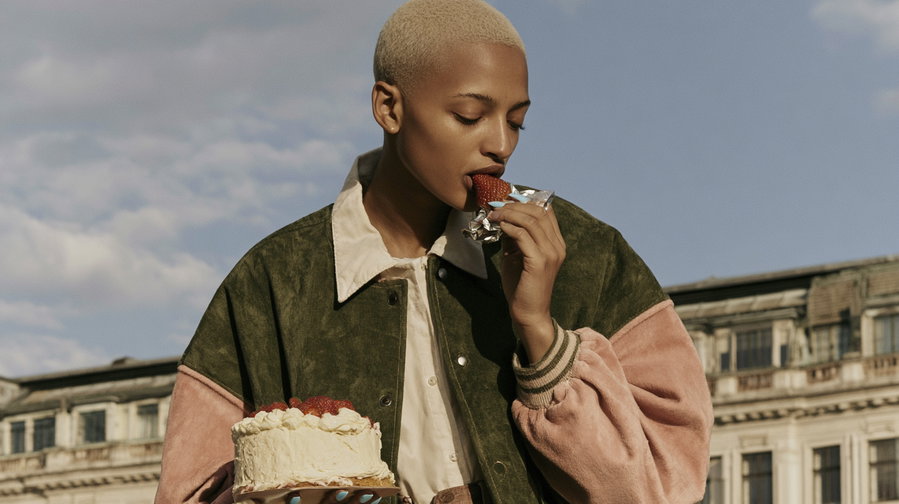 A close-up of a person with short blonde hair and dark skin, wearing a green and pink letterman jacket, holding a cake with white frosting and strawberries in one hand, and eating a strawberry from a wrapper with the other. The background features a building with multiple windows and a partly cloudy sky.