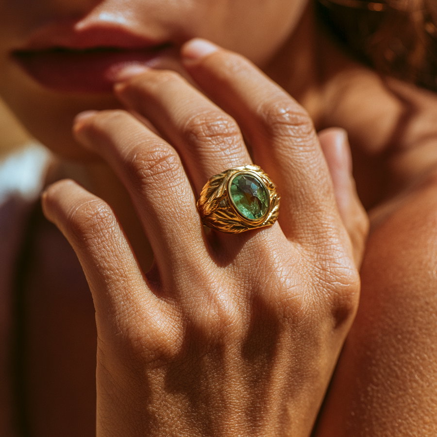 A person's hand with a gold leaf-shaped ring and green gemstone, softly blurred background