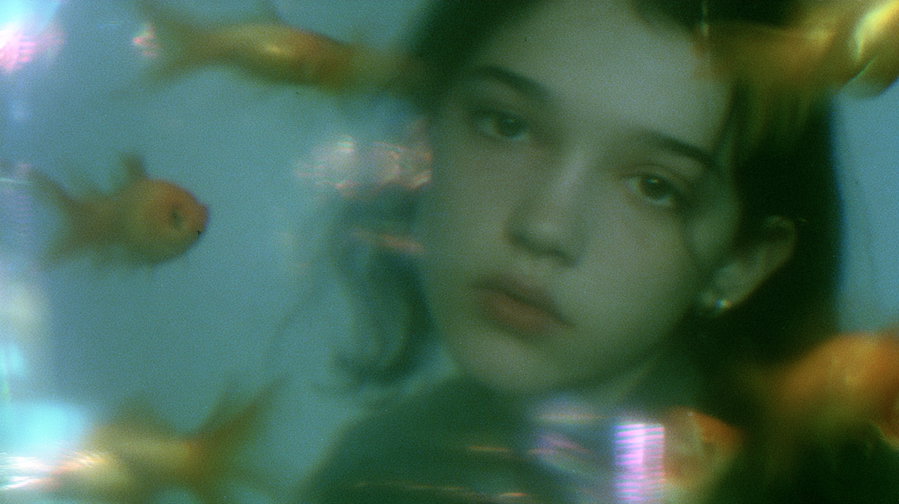 A close-up portrait of a person behind a goldfish tank, face distorted by water ripples and reflections. Goldfish swim throughout the scene, creating a surreal ambiance. The background features an empty room with a nostalgic, low-resolution look, using cool tones and soft light.