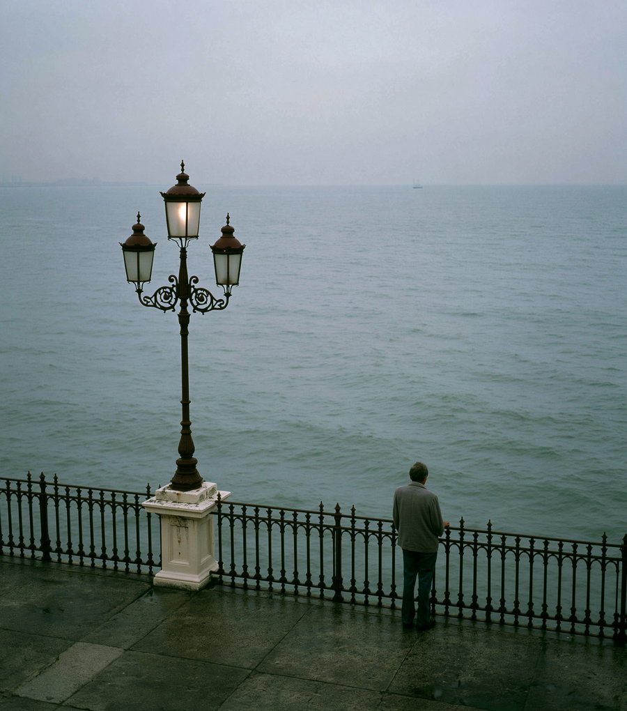 A man standing alone by a wet railing overlooking the calm sea under a cloudy sky, with a street lamp nearby.