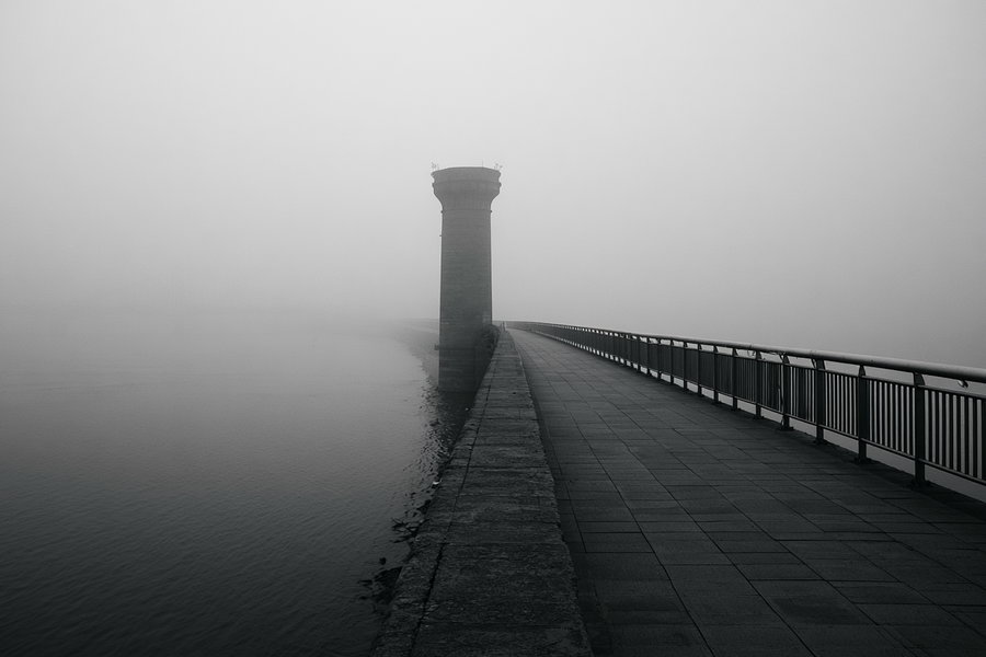A foggy black and white photograph of a walkway along the Han River, featuring an old bridge pillar in the distance, with soft mist surrounding the scene and a minimal composition.