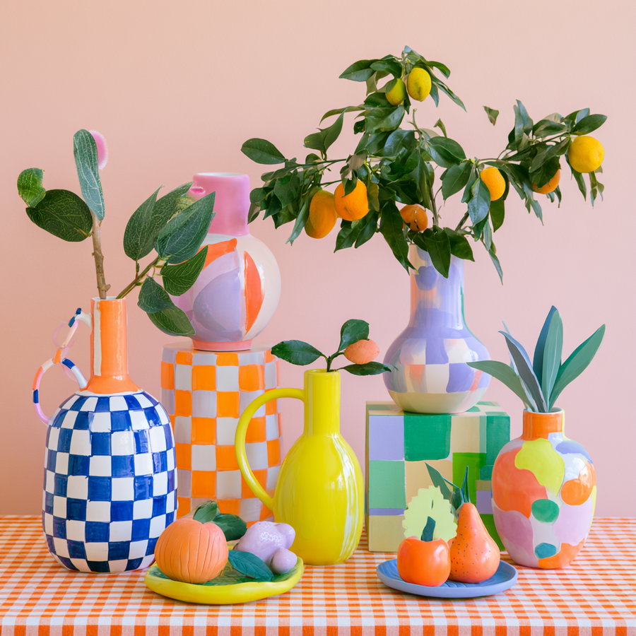 A lively arrangement of colorful vases in bright neon and pastel shades on a gingham tablecloth. The vases are various patterns, including checks and solid colors, and contain fresh flowers and fruits.