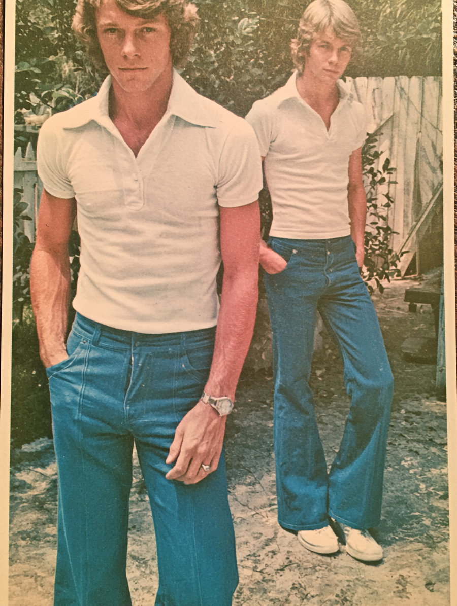Two males dressed in 1970s fashion; one in a white polo shirt and blue pants, the other in a similar outfit, standing against a natural background.
