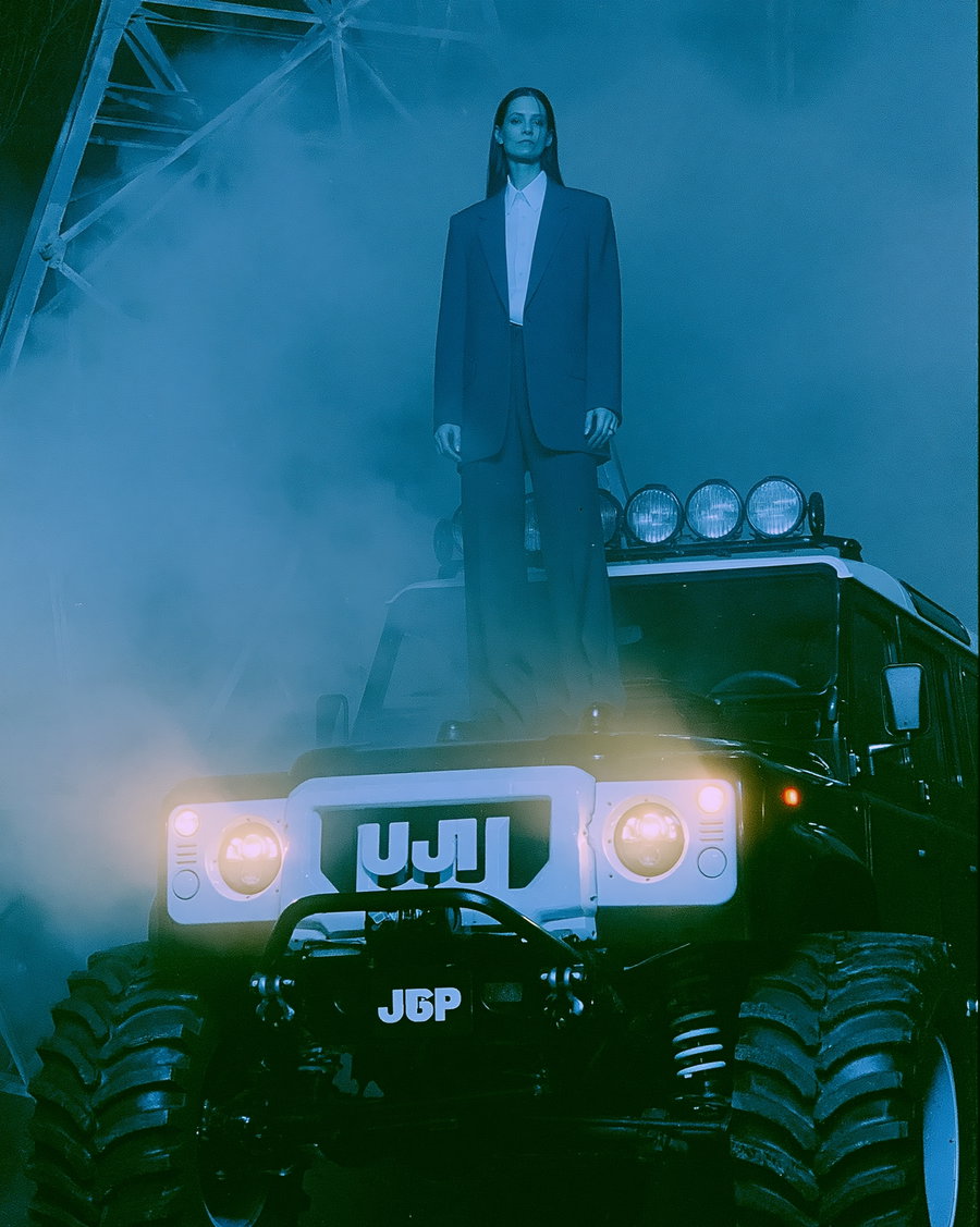 A woman wearing a black suit stands atop a black SUV, surrounded by fog and dramatic lighting.