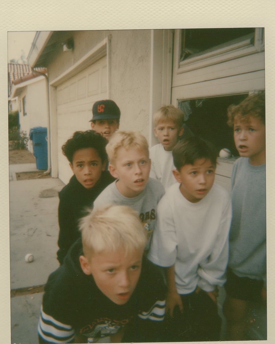 Group of boys with varied hairstyles, looking shocked beside a shattered garage window. A baseball lies on the ground nearby.