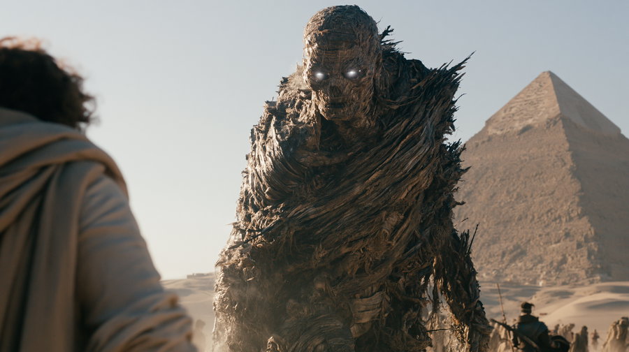 A giant, intricately designed mummy stands in the Egyptian desert, with a pyramid visible in the background under a clear sky.