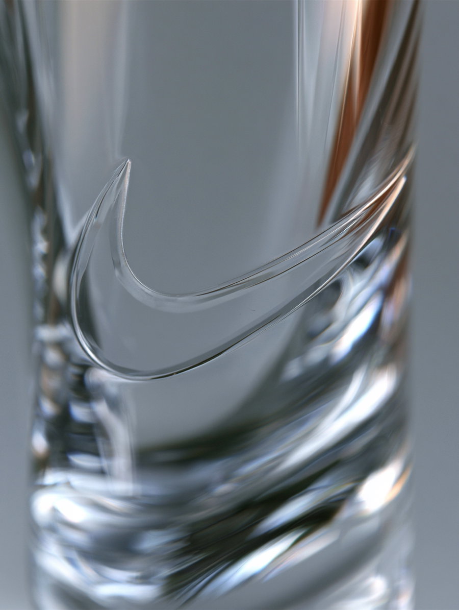 A transparent crystal object with an engraved swoosh logo, showing light reflections and a colorless finish.
