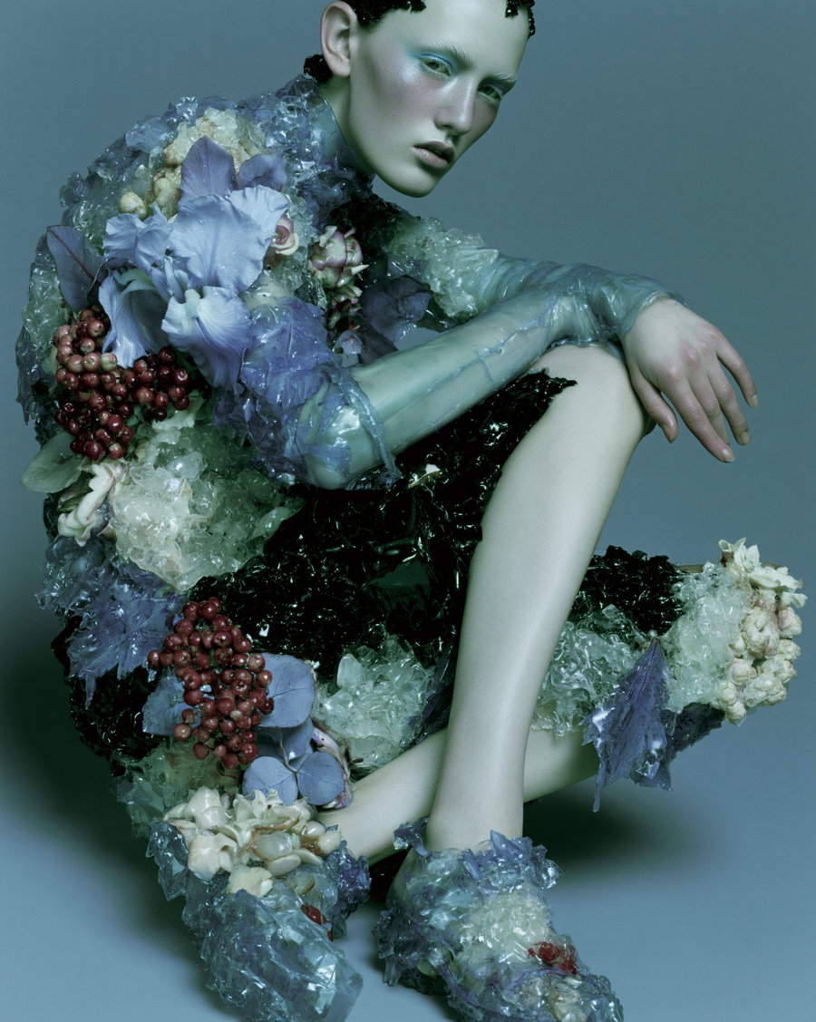 A semi-reclining figure adorns a dress made of translucent gel, flowers, and crystals, set against a blue metal background, with cold clinical lighting.