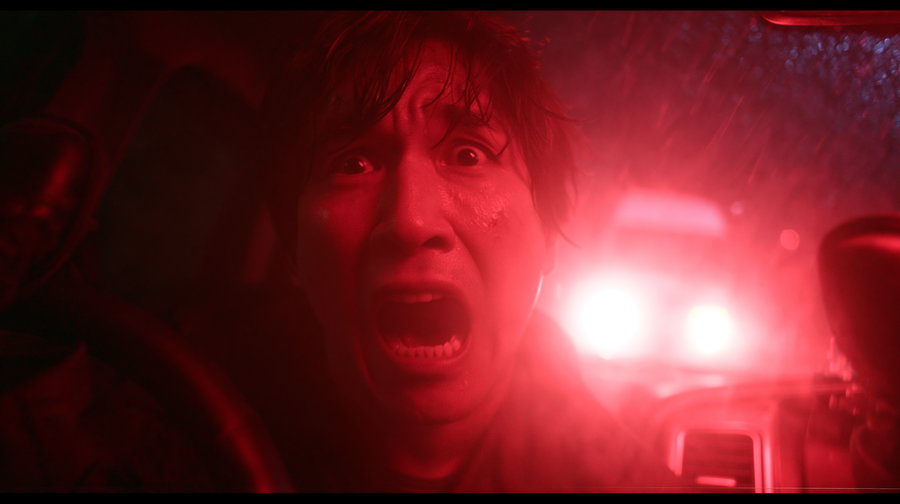 A terrified man in a dark-lit SUV, kicking a car window, illuminated by red light and truck headlights through a rain-streaked windshield.